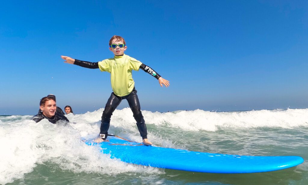 a boy rides a wave with the help of a qualified campsurf instructor on a blue bird sunny day